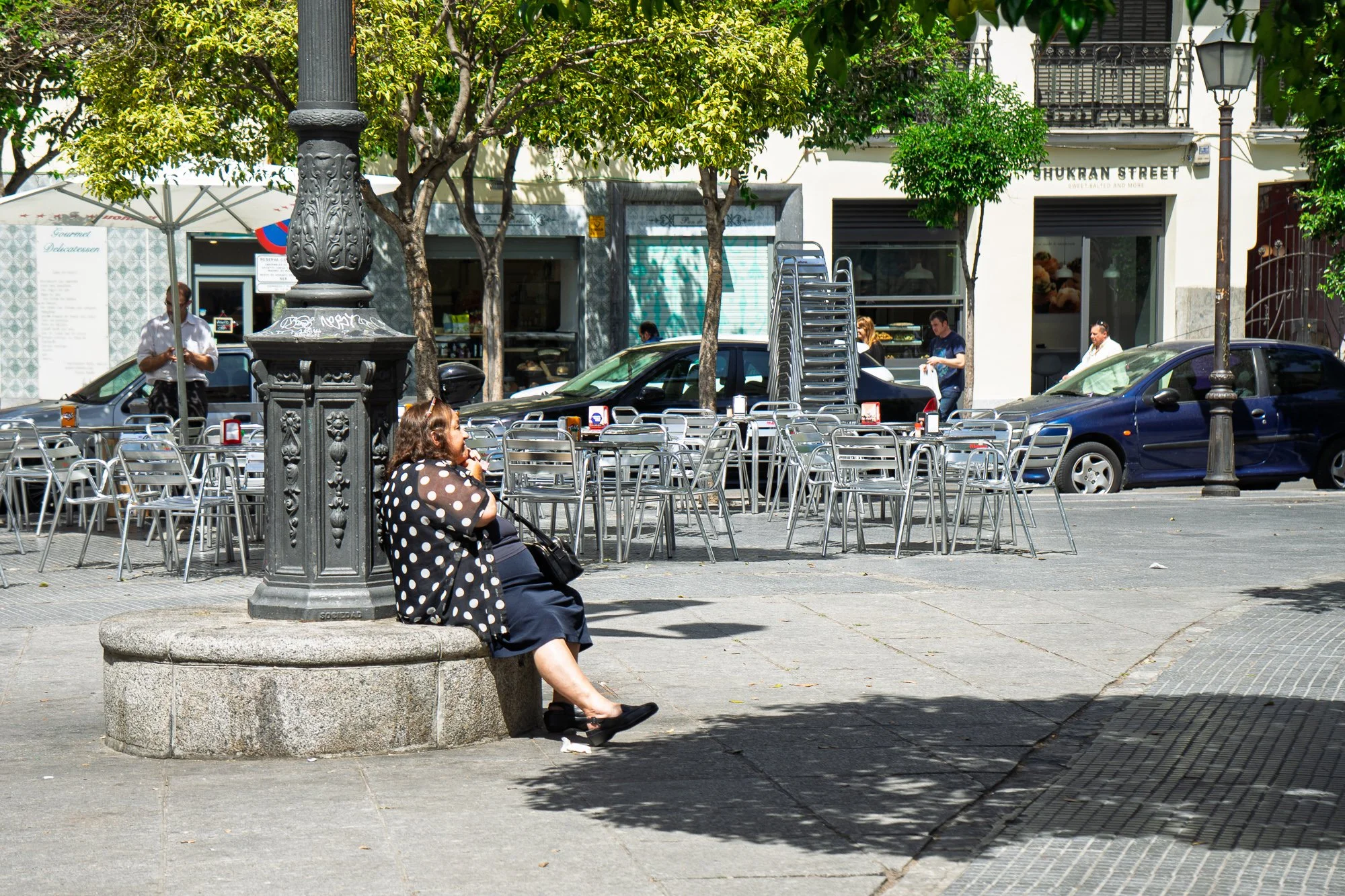 A woman in a polka-dot blouse rests against a cast-iron lamppost base in the shade, empty café chairs behind her