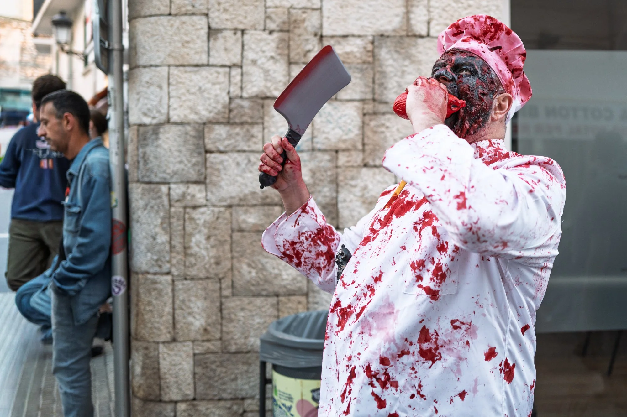 Zombie chef in a blood-soaked white jacket raises a cleaver and bites into a heart by a stone wall, Sitges — Leica Q3