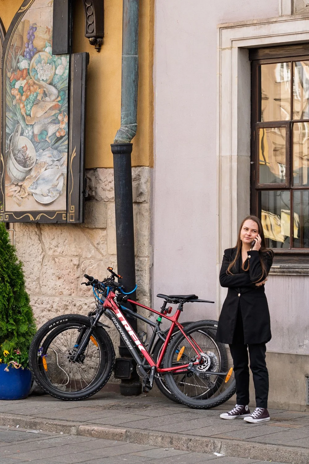 Woman in a black blazer on the phone beside bikes and a painted shop sign, Warsaw Old Town — Sony a7R IV