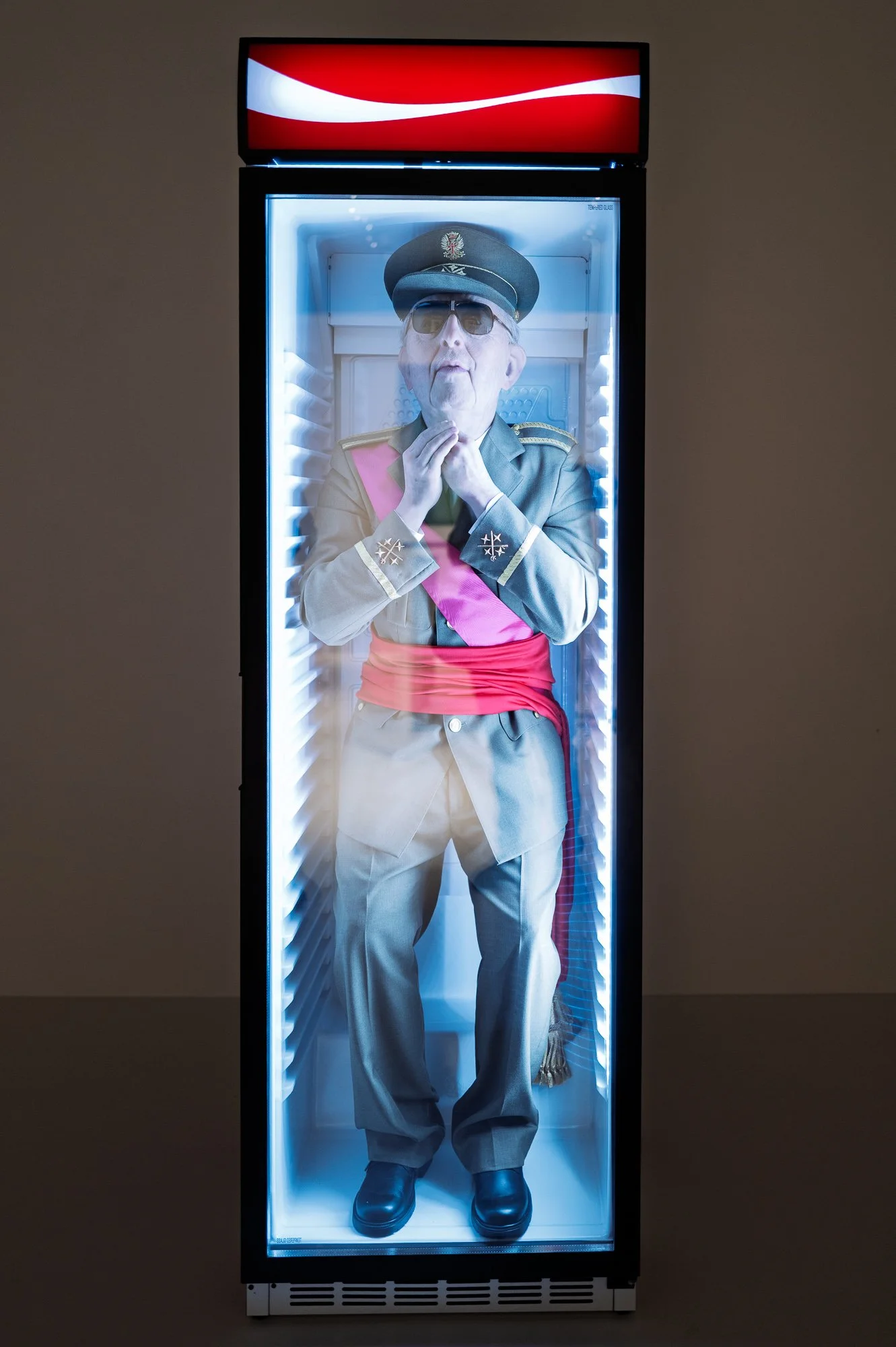 A lifelike figure of Franco in uniform and sunglasses standing inside a lit drinks fridge with a red and white sign on top