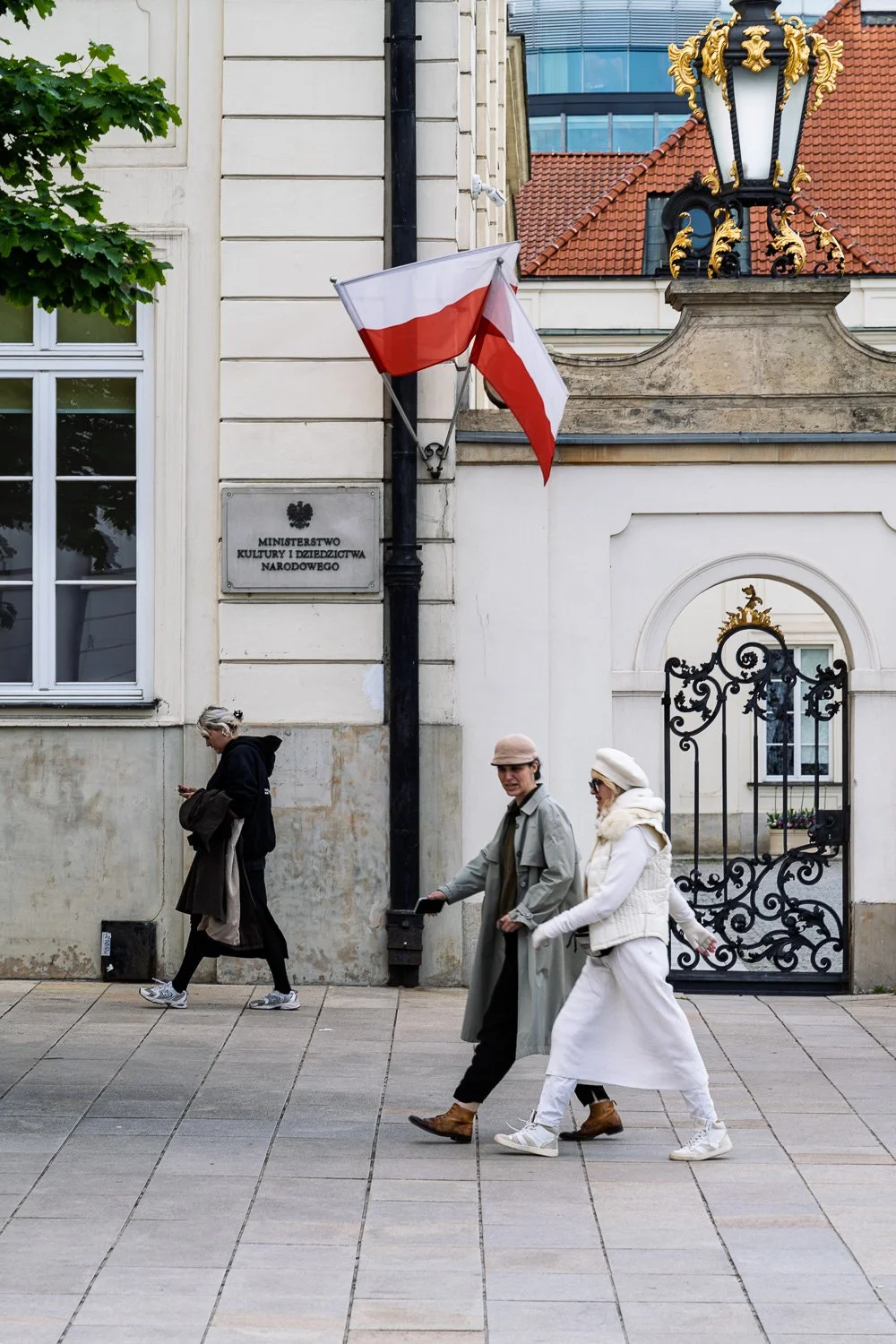 Three women walk past the Ministry of Culture sign, Polish flags and a gilded lantern, Warsaw — Sony a7R IV