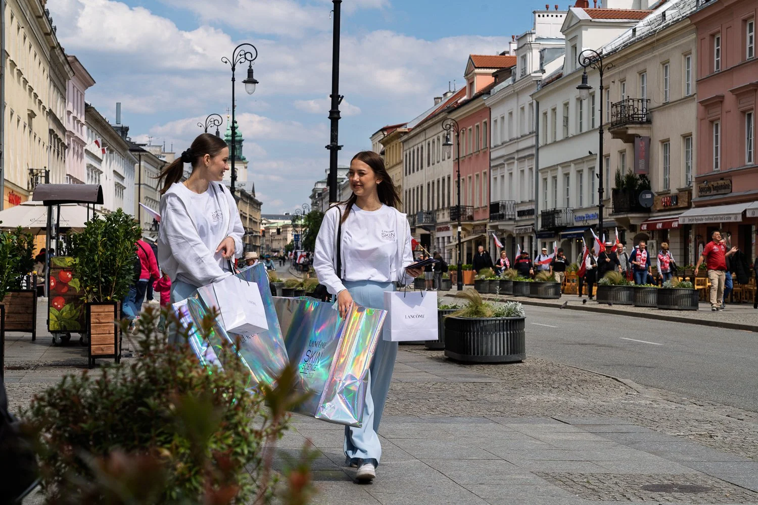 Two young women carry holographic Lancôme bags as a march with flags passes behind, Warsaw — Sony a7R IV