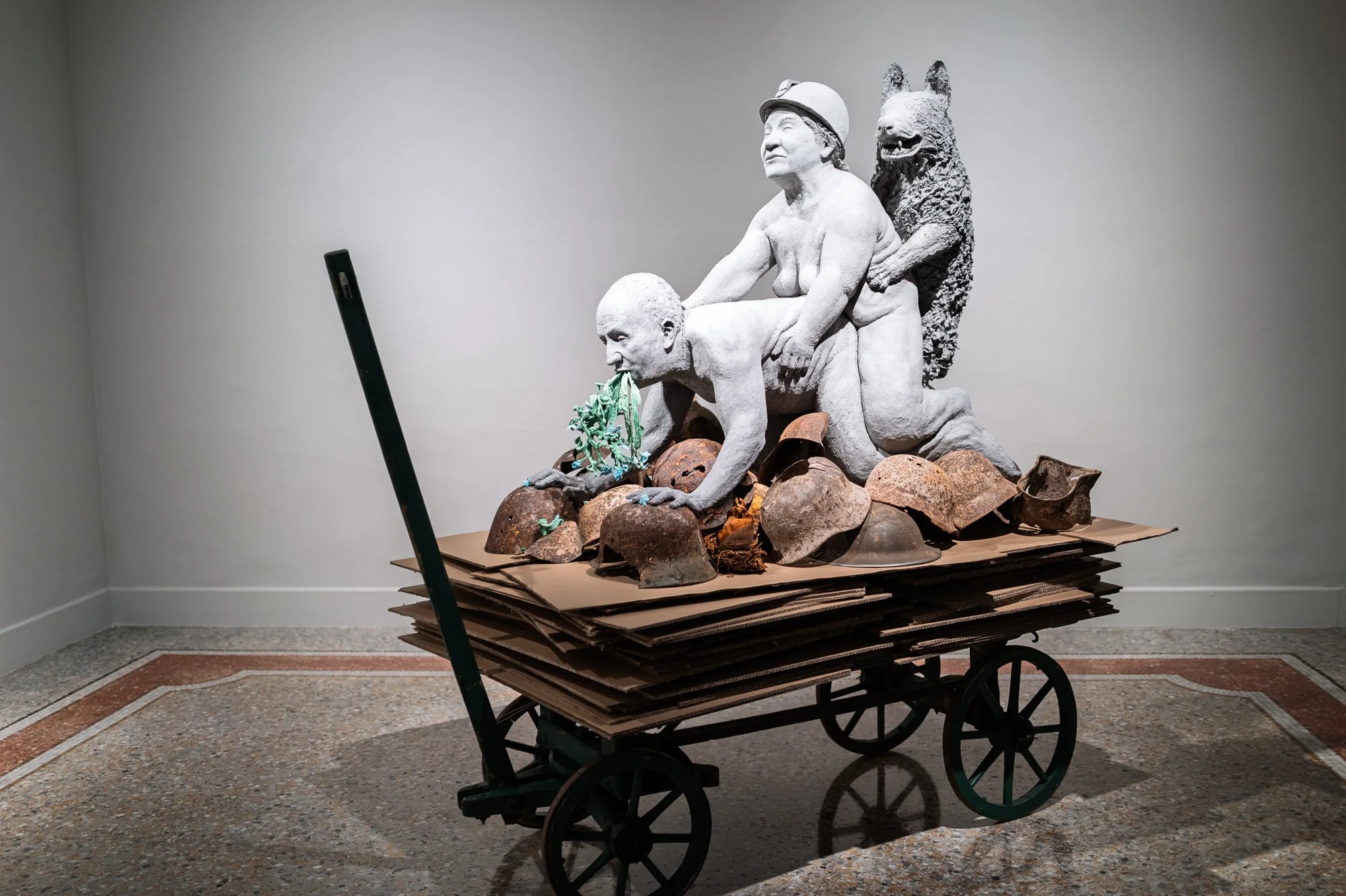 Sculpture on a wooden handcart: a figure on all fours spitting blue flowers, a woman in a miner's helmet and a dog, on rusty helmets