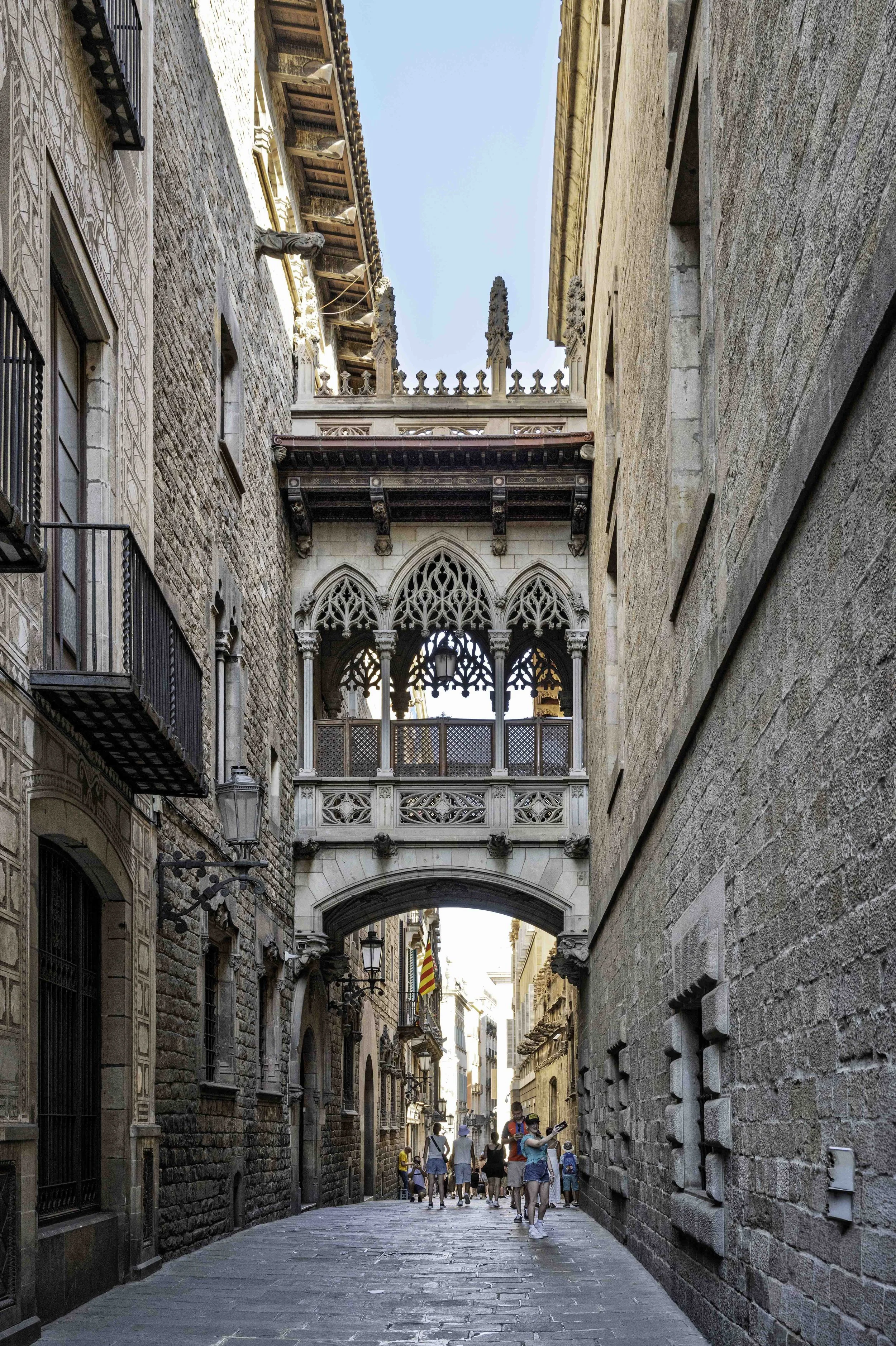 Gothic Quarter Barcelona street view looking up at the ornate Pont del Bisbe bridge with pedestrians walking below through the narrow medieval alley — Leica Q3