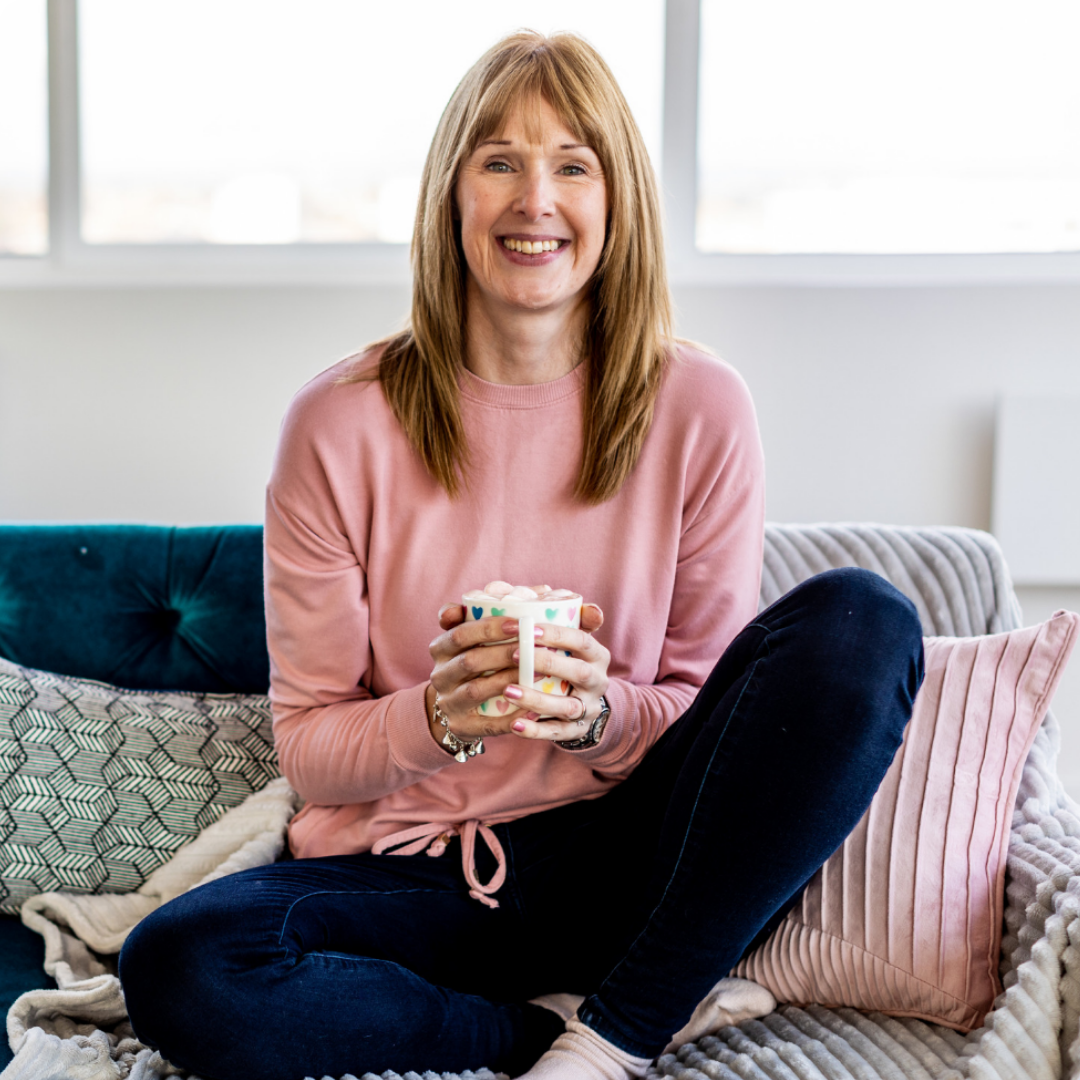 A woman sitting on a couch with pillows, holding a mug, smiling, wearing a pink sweatshirt and dark pants, in a bright room.