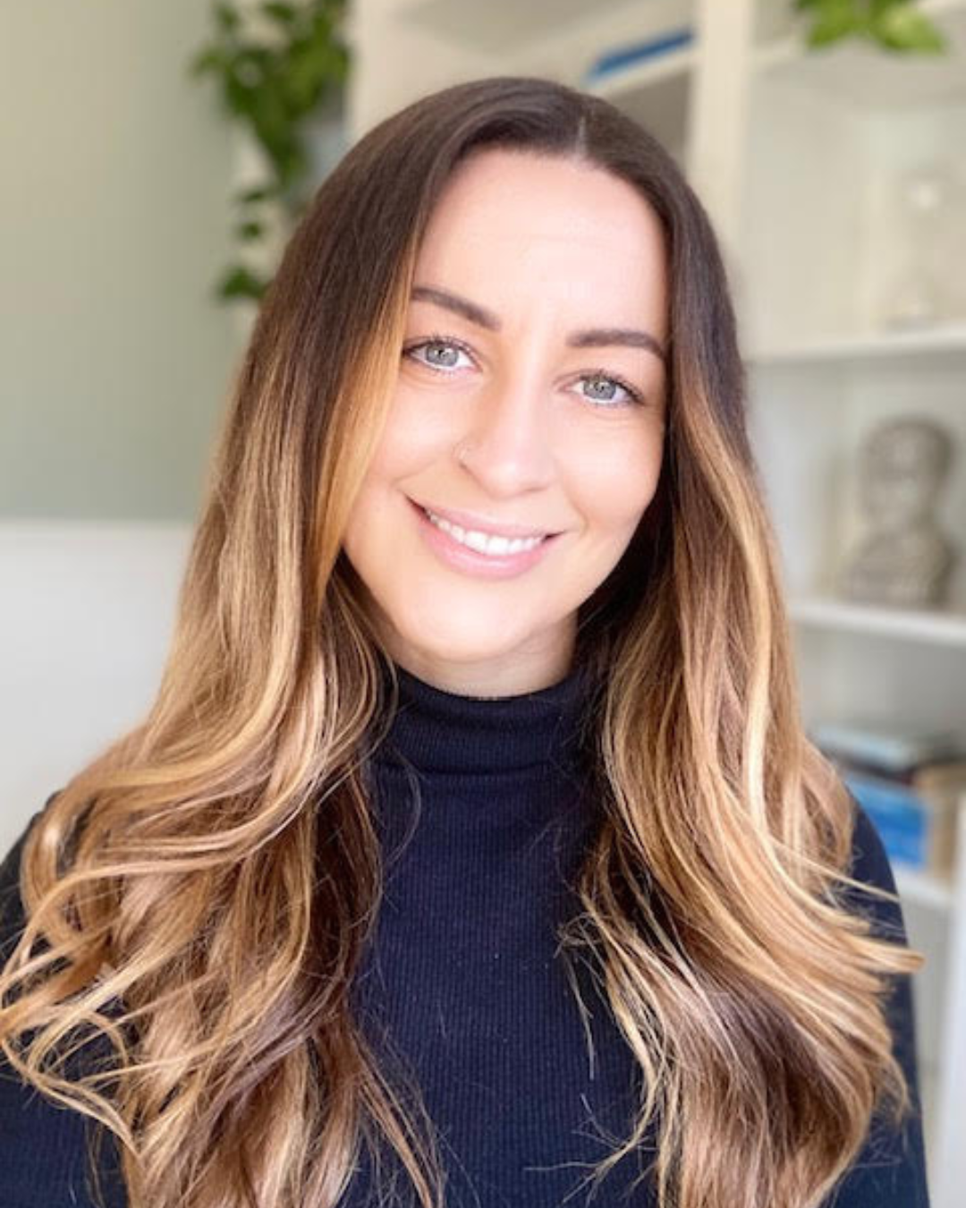 A woman with long, wavy, ombré hair smiling at the camera, wearing a dark turtleneck, in an indoor setting with blurred background shelves and green plants.