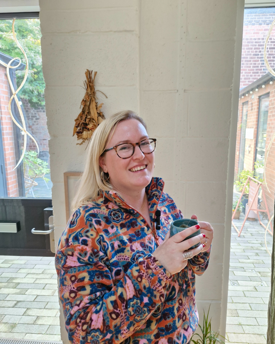 A woman with blonde hair and glasses smiling, holding a mug, standing indoors near a window with a view of a brick patio and greenery.
