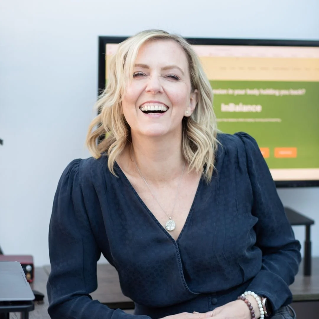 A smiling blonde woman with shoulder-length hair, wearing a navy blue blouse and jewelry, sitting in an office environment with a computer screen and desk in the background.