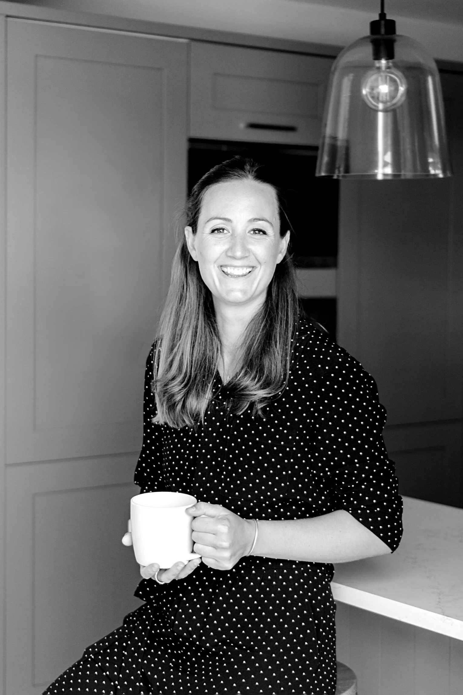 A woman with long hair, smiling, holding a mug, sitting in a kitchen with modern cabinets and a hanging lamp.