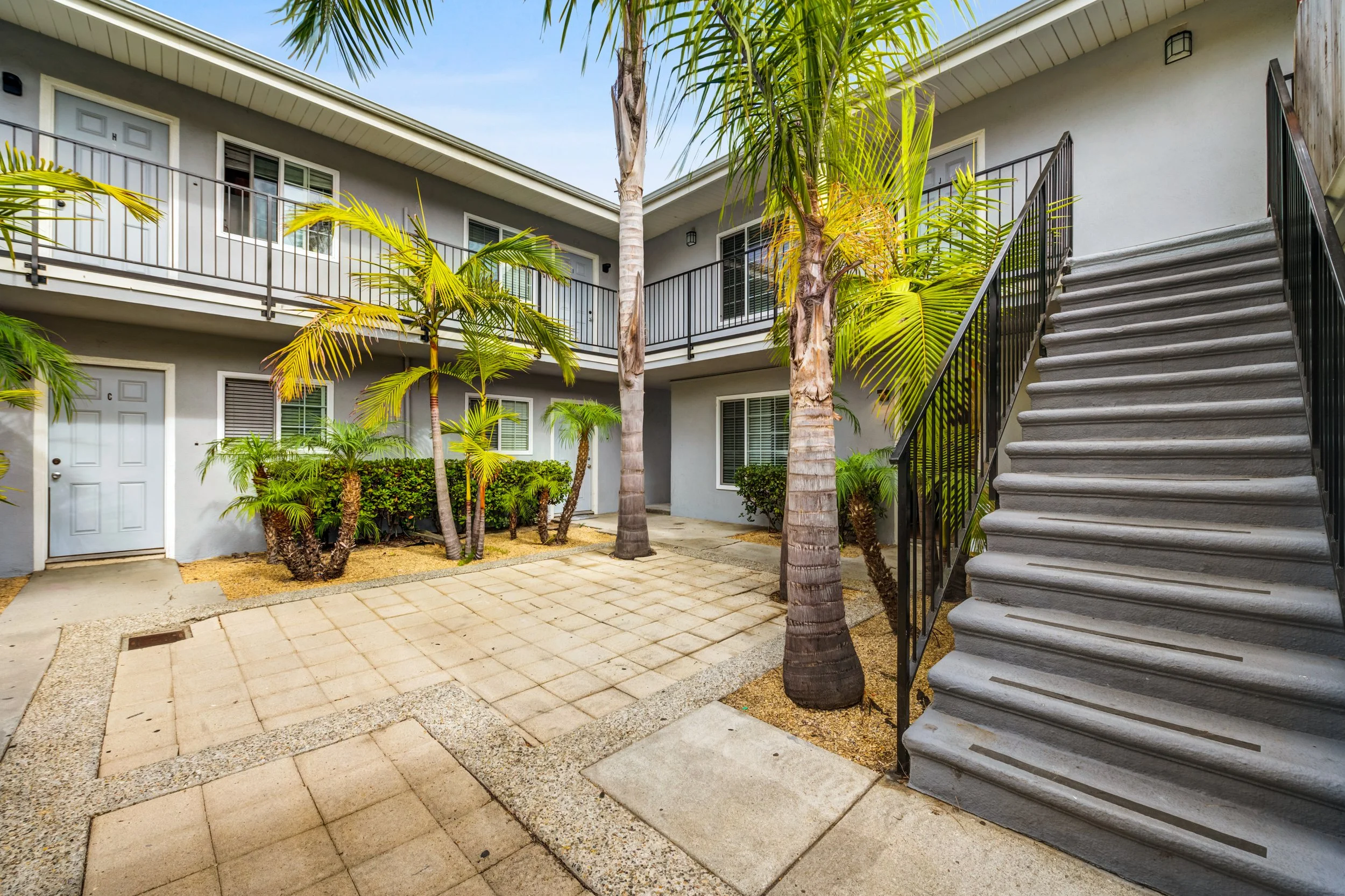 Exterior view of a two-story apartment building with gray walls, black railings, and a staircase on the right. There are palm trees and green shrubs in the courtyard area.
