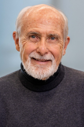A smiling elderly man with white hair and a beard, wearing a dark sweater, against a light blue background.