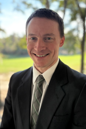 A man in a dark suit, white shirt, and patterned tie smiling outdoors with trees and greenery in the background.