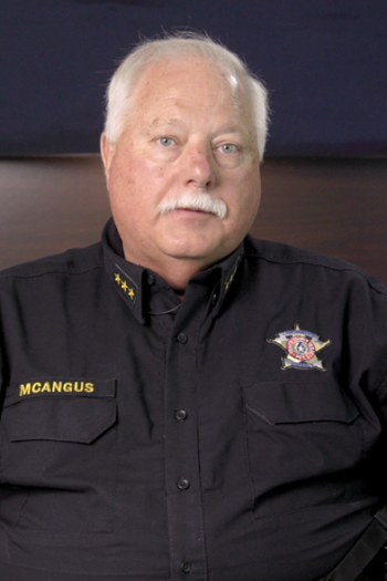 A middle-aged man with white hair and a mustache, wearing a black uniform shirt with a badge and the name tag 'MCANGUS'.