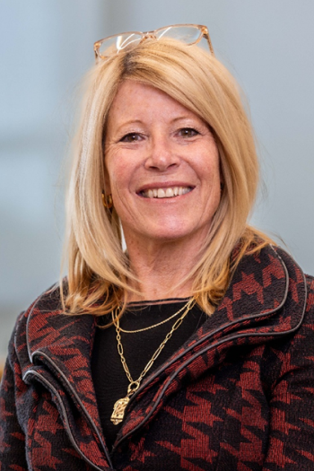 A middle-aged woman with blonde hair smiling, wearing glasses on her head, gold earrings, a gold necklace, and a patterned jacket.