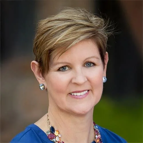 A woman with short blonde hair wearing a blue top, pearl earrings, and a colorful necklace, smiling outdoors.