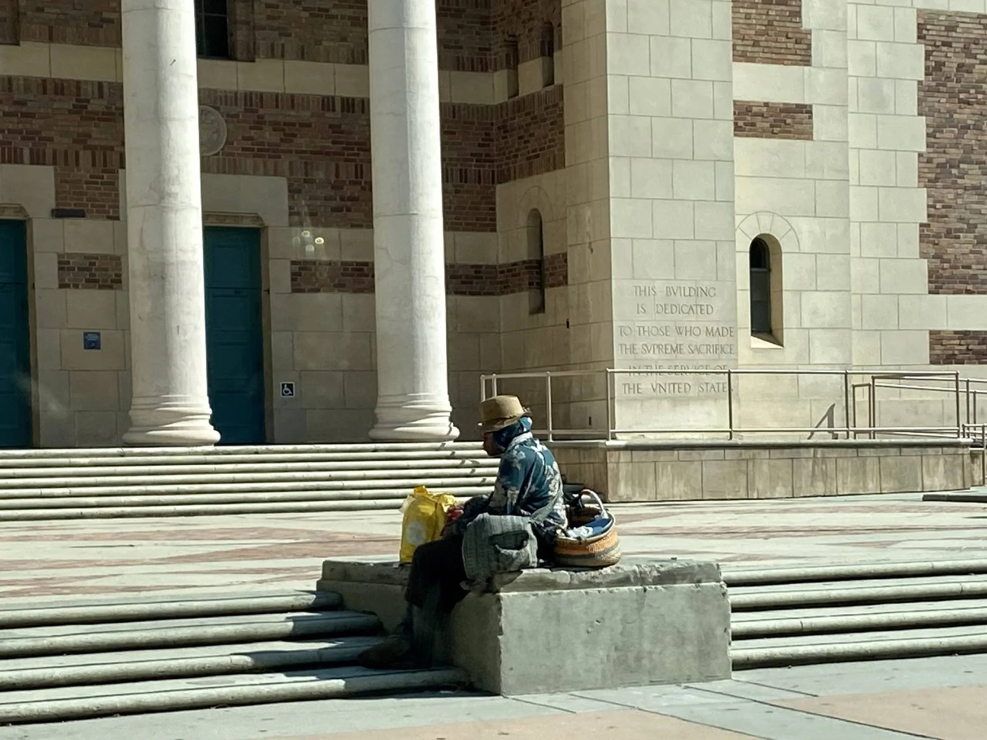 A person sitting on a concrete block outside a building with large white columns, holding a yellow bag and surrounded by backpacks and belongings.