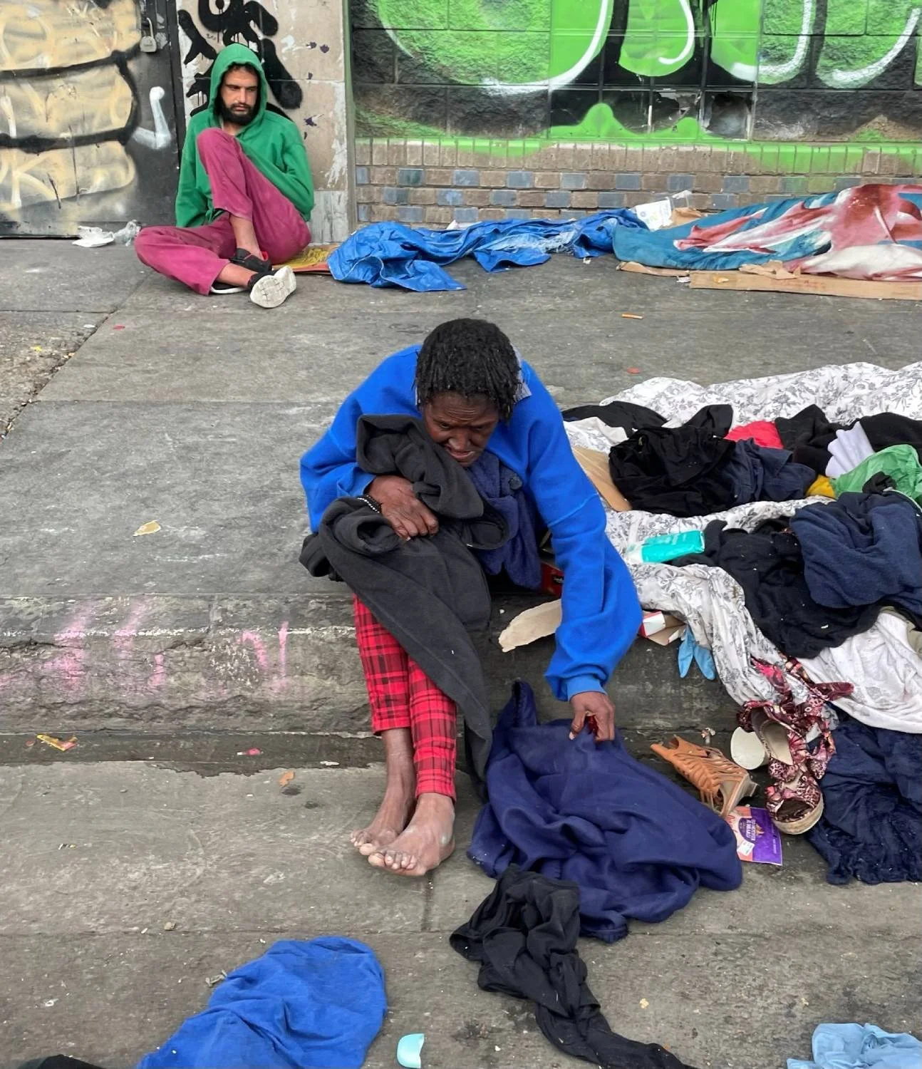 Two people sitting on a city sidewalk with scattered clothing and belongings. One person in a blue jacket and red plaid pants is leaning forward, holding a black item and reaching into a bag. A second individual in a green hoodie and maroon pants sits against a wall in the background.