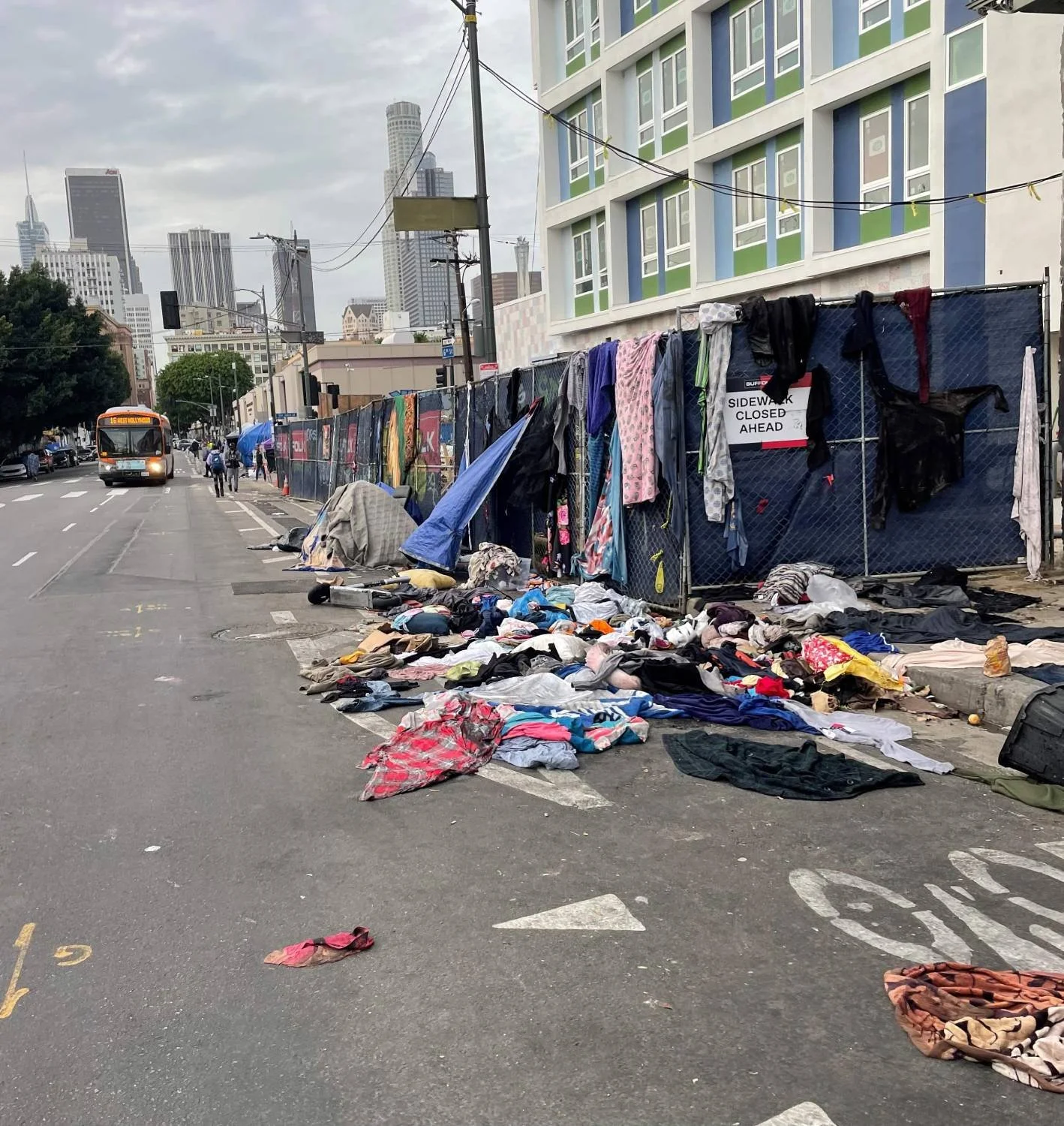 Street scene with overturned clothing, bags, and personal items on the sidewalk, behind a construction fence, with a city skyline featuring tall buildings and a bus in the background.