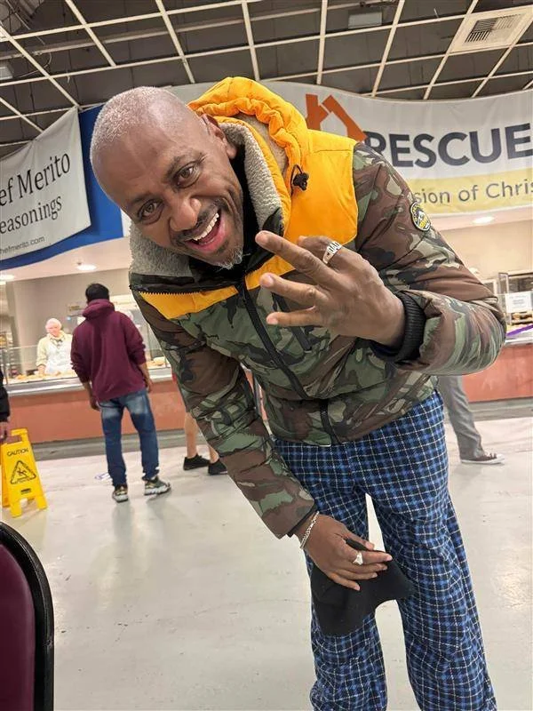 A smiling man making a peace sign with his right hand, wearing a camouflage jacket with a yellow hood, blue plaid pants, and jewelry, in an indoor public space.