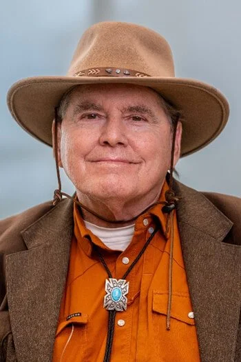 Older man wearing a tan cowboy hat, brown jacket, orange shirt, with a silver sheriff badge necklace, smiling.
