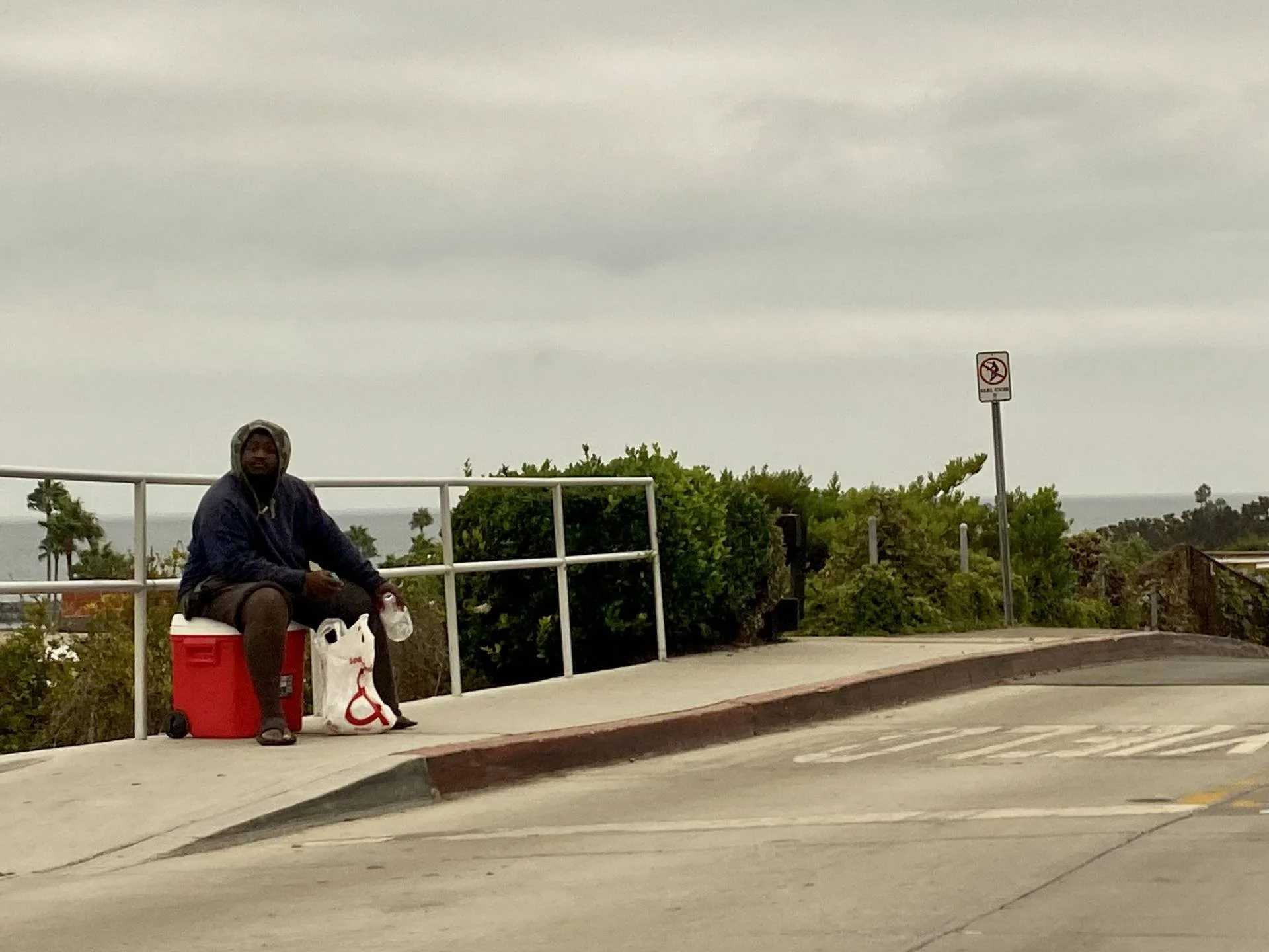 A man sitting on a red cooler on the sidewalk, wearing a hooded jacket and sandals, holding a plastic bottle and a bag with grocery items, near greenery and a no-smoking sign under a cloudy sky.