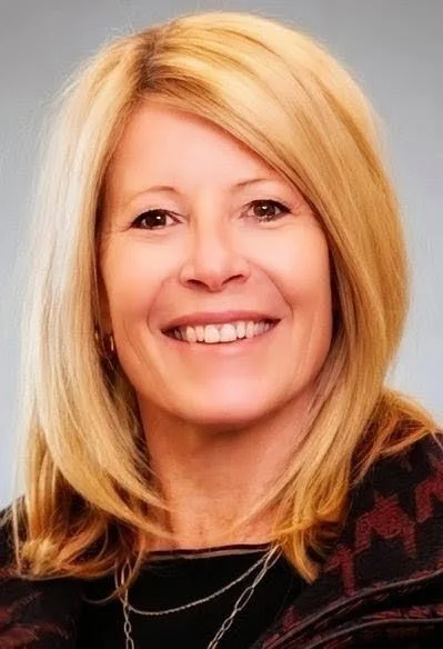 A middle-aged woman with blonde hair smiling, wearing glasses on her head, gold earrings, a gold necklace, and a patterned jacket.