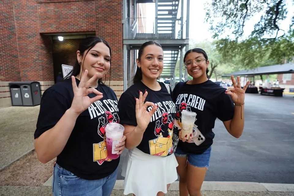 Three young women smiling and making the 'OK' hand gesture outdoors, wearing matching black t-shirts with a graphic design, one holding a drink, in front of a brick building and a staircase.