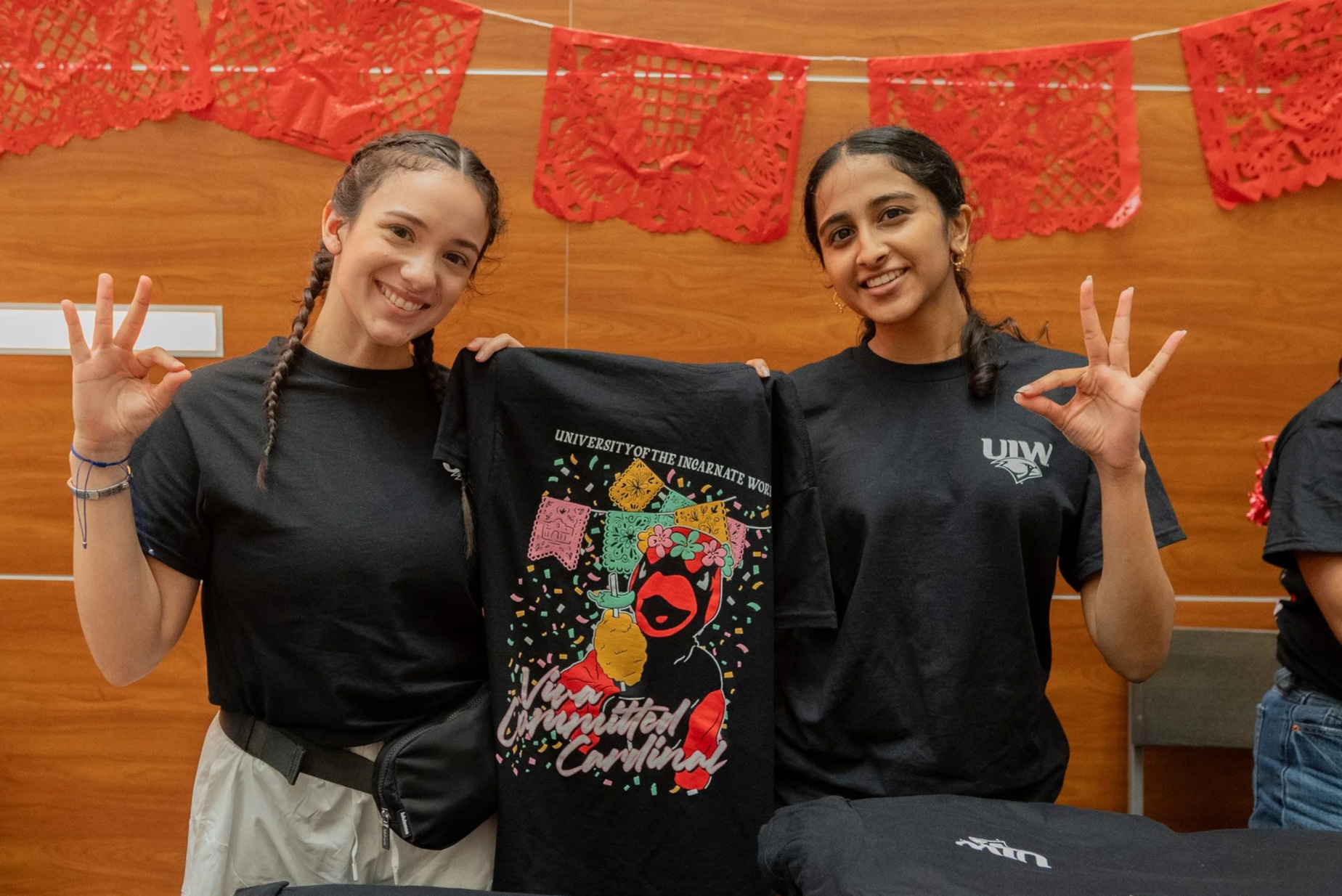 Two young women smiling and making 'OK' hand gestures, holding a black backpack with colorful graphics and text that reads 'University of the Incarnate Word' and 'Viva La Communidad Cardinal,' standing in front of a wooden wall decorated with red paper banners.