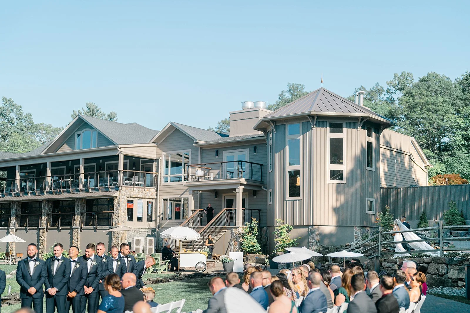 A wedding ceremony taking place outdoors in front of a modern, multi-story house with a deck. The bride and groom are walking towards the house, surrounded by seated guests and groomsmen dressed in suits.