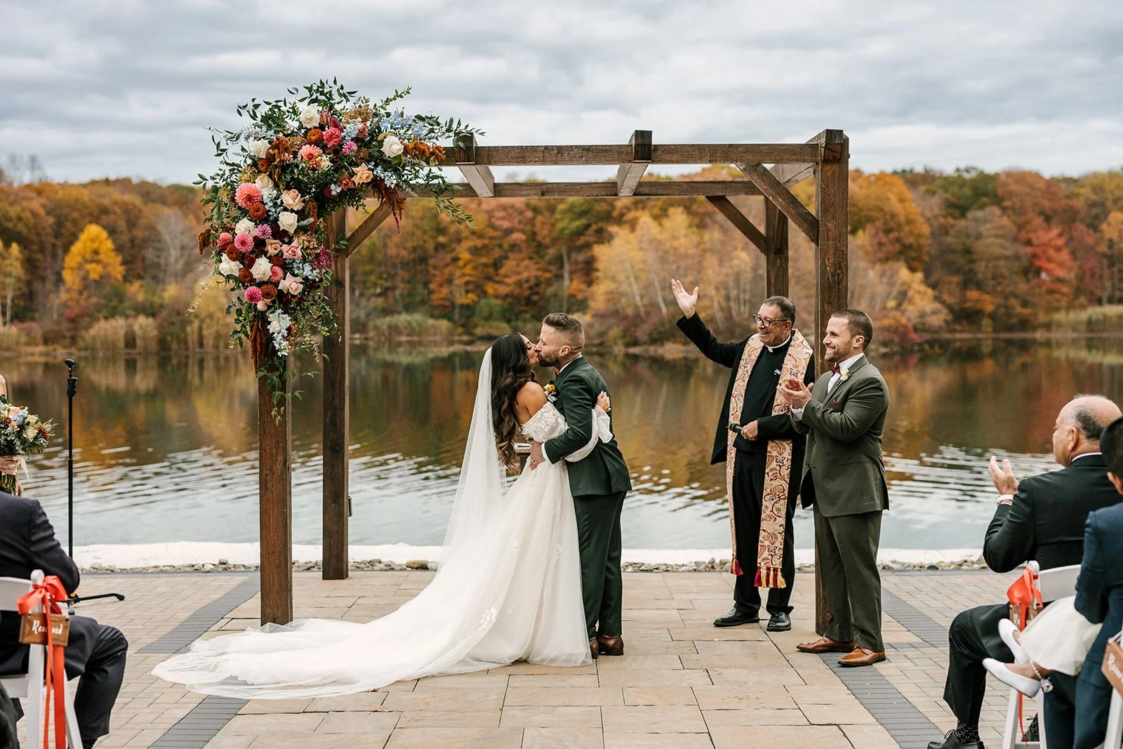 Bride and groom kissing during their outdoor wedding ceremony by a lake with fall foliage in the background. An officiant and guest are clapping, with chairs decorated with orange bows.