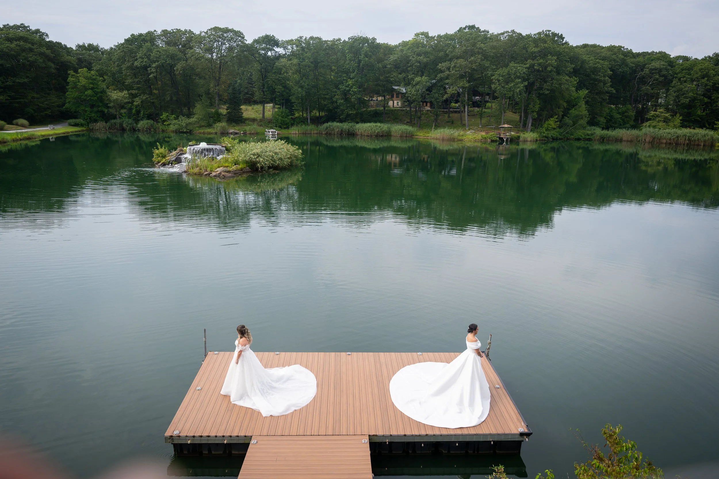Two brides in white wedding dresses standing on a wooden dock by a calm lake, with lush green trees in the background.