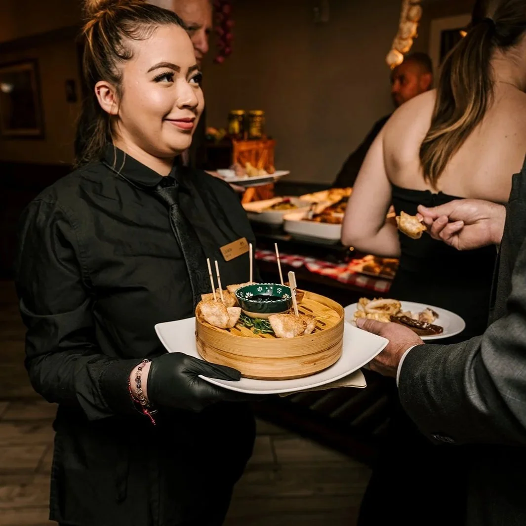 A waitress with a name tag serving food in a restaurant, holding a tray with assorted appetizers, while a customer reaches for food in the background.