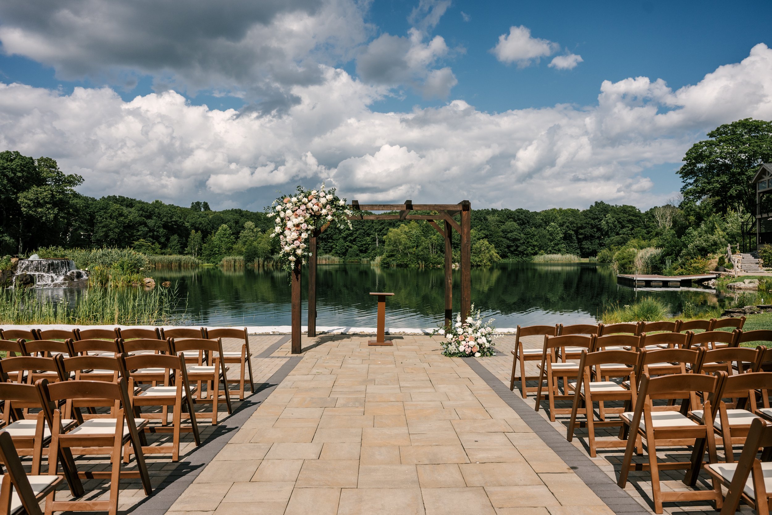 Lakeside wedding ceremony setup outdoors by a lake, with rows of chairs facing a wooden arch decorated with flowers, surrounded by lush greenery and a partly cloudy sky.