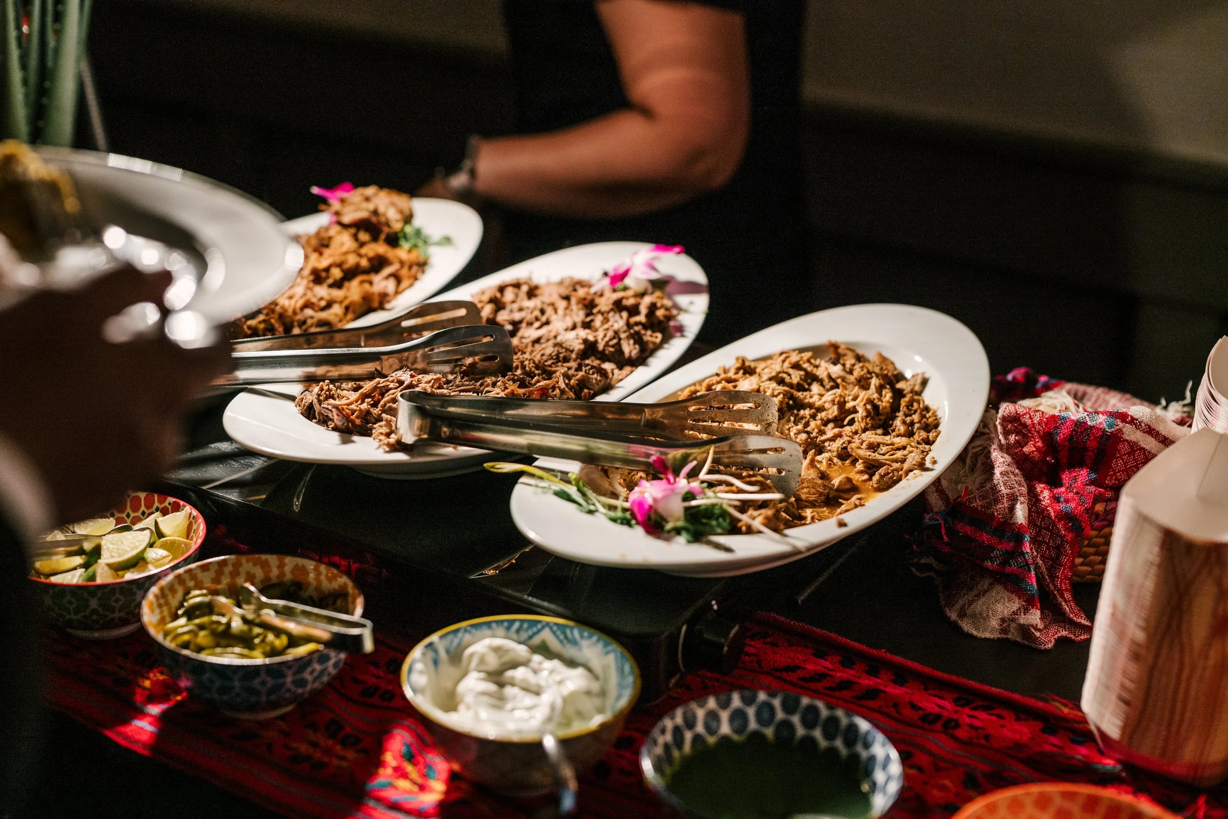 Plates of shredded beef or pork, garnished with flowers, set on a table with bowls of lime slices, pickles, and sauces, with a red patterned tablecloth.