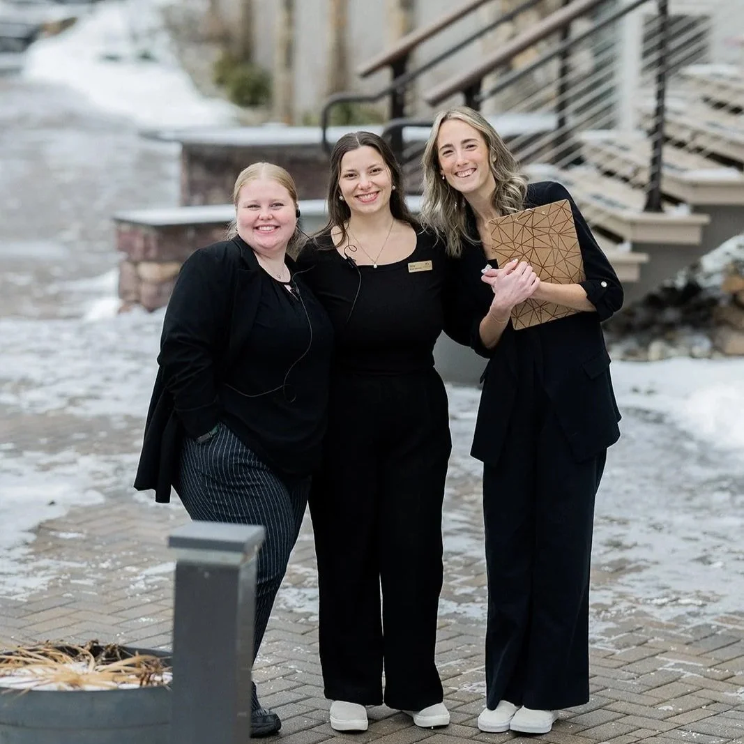 Three women standing outside on a snowy brick walkway, smiling at the camera, with stairs and a building in the background.