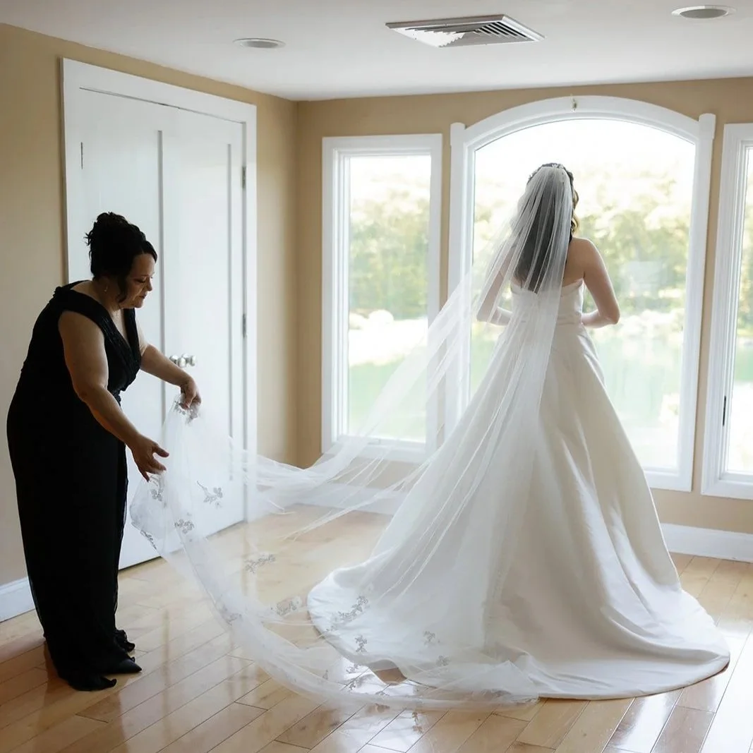 A bride in a white wedding gown with a long veil stands by large windows, while a woman helping her with the veil in a black dress.