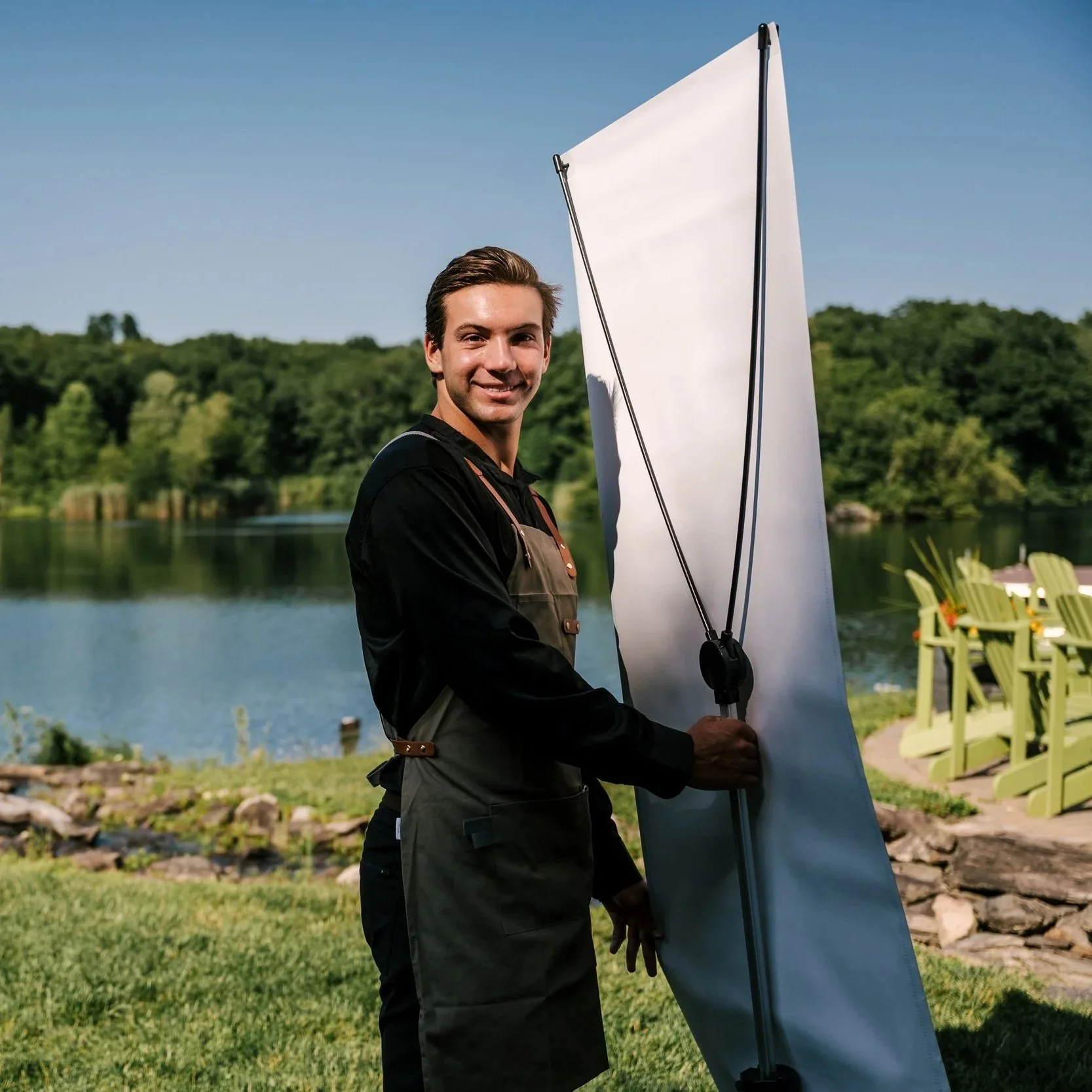 A smiling man holding a large white sail or kite outdoors near a lake, with green trees and yellow chairs in the background.