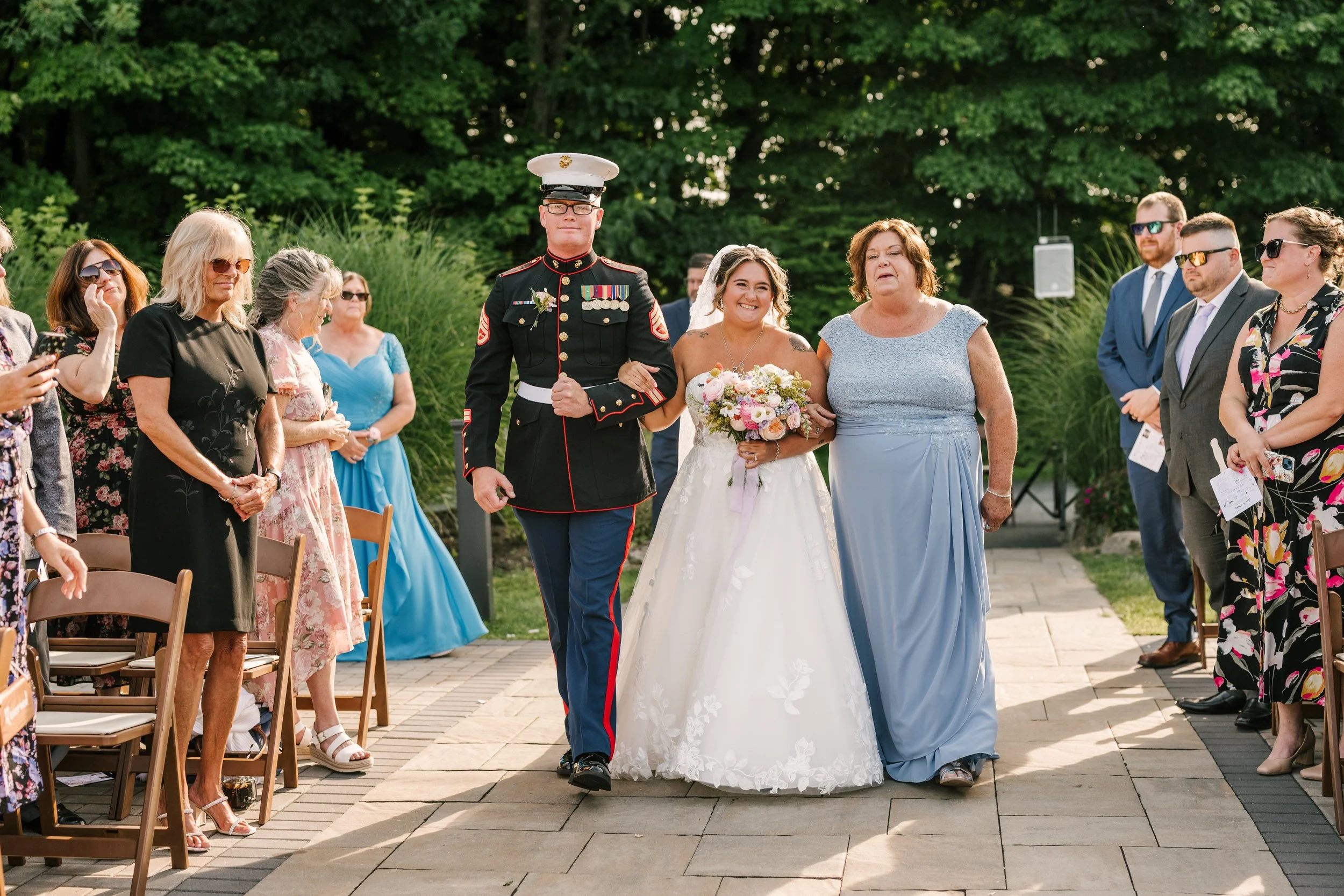 Diverse guests attending a wedding ceremony outdoors, with some women wearing sunglasses and floral dresses and a military officer in uniform walking down the aisle with a bride holding a bouquet of flowers.