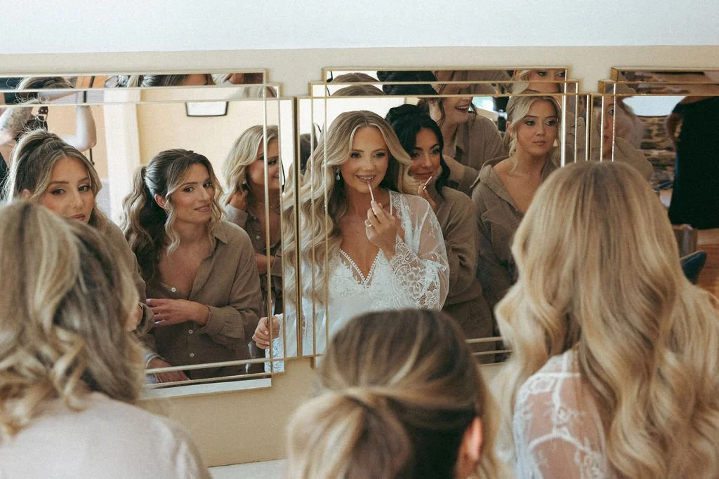 Several women, including a bride in a white lace robe, look at her reflection in a mirror while getting ready for a wedding.