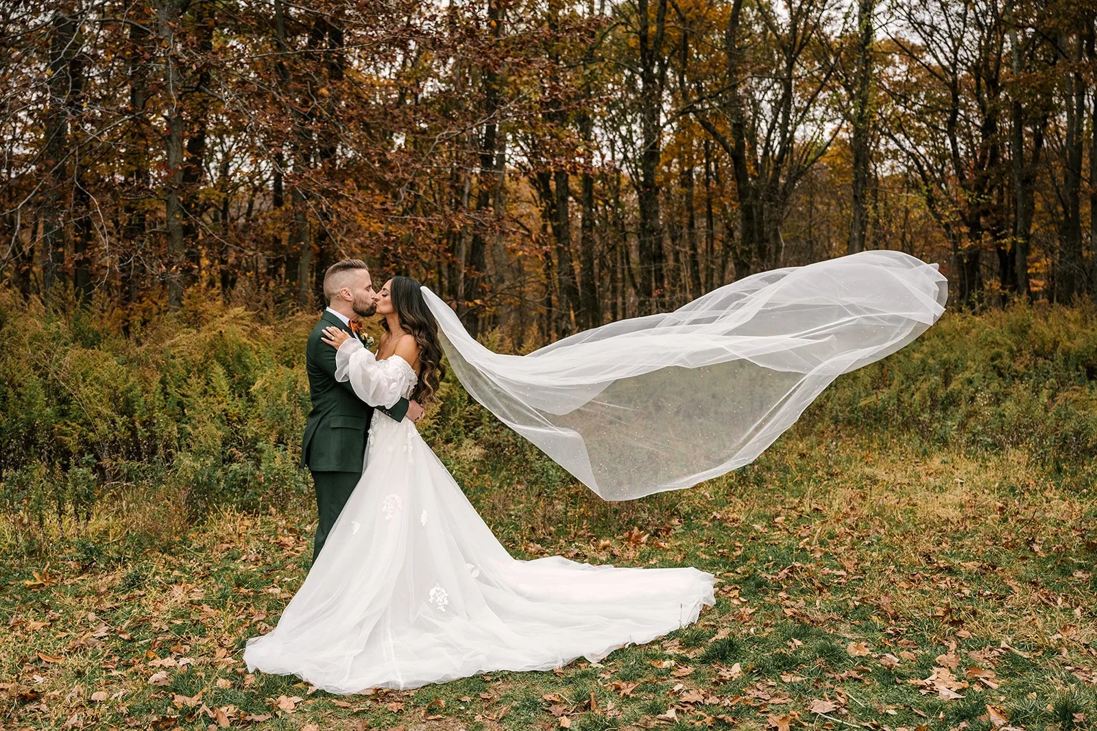 Bride and groom in wedding attire sharing a kiss outdoors during autumn with fallen leaves, woman has flowing veil.