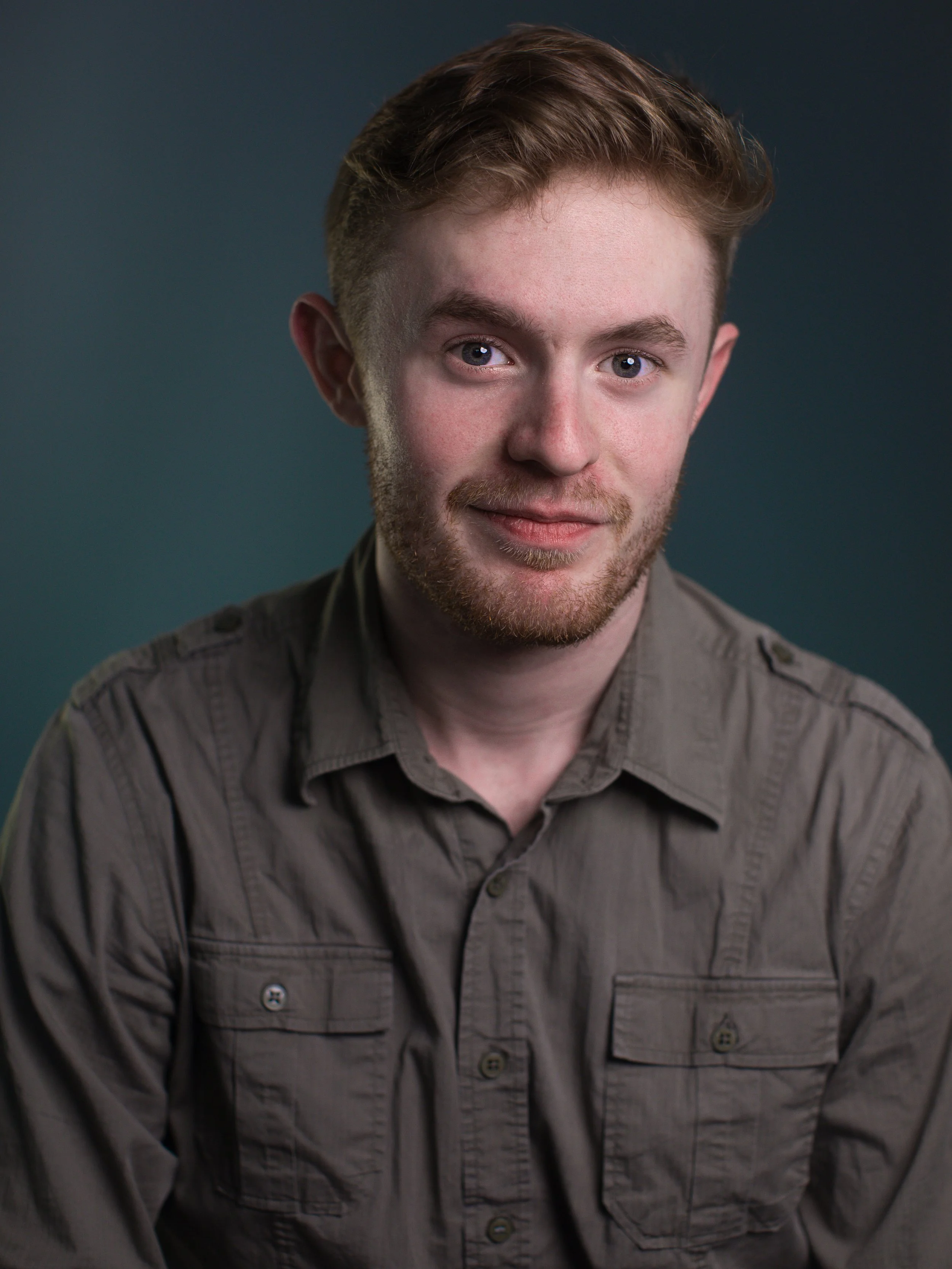A young man with light brown hair and a beard wearing an olive-green button-up shirt. He is smiling slightly and looking at the camera, with a neutral dark background.