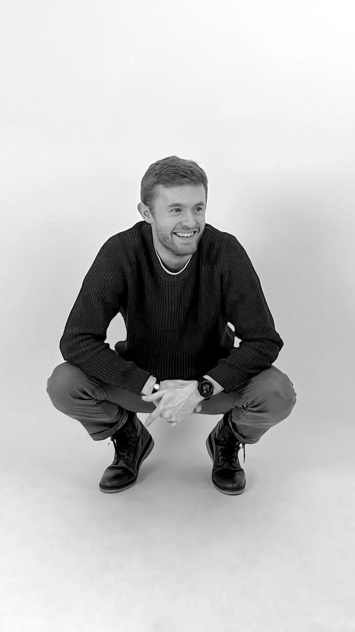 A man crouching and smiling, wearing a black sweater, khaki pants, and black boots, against a plain white background, black and white photo.
