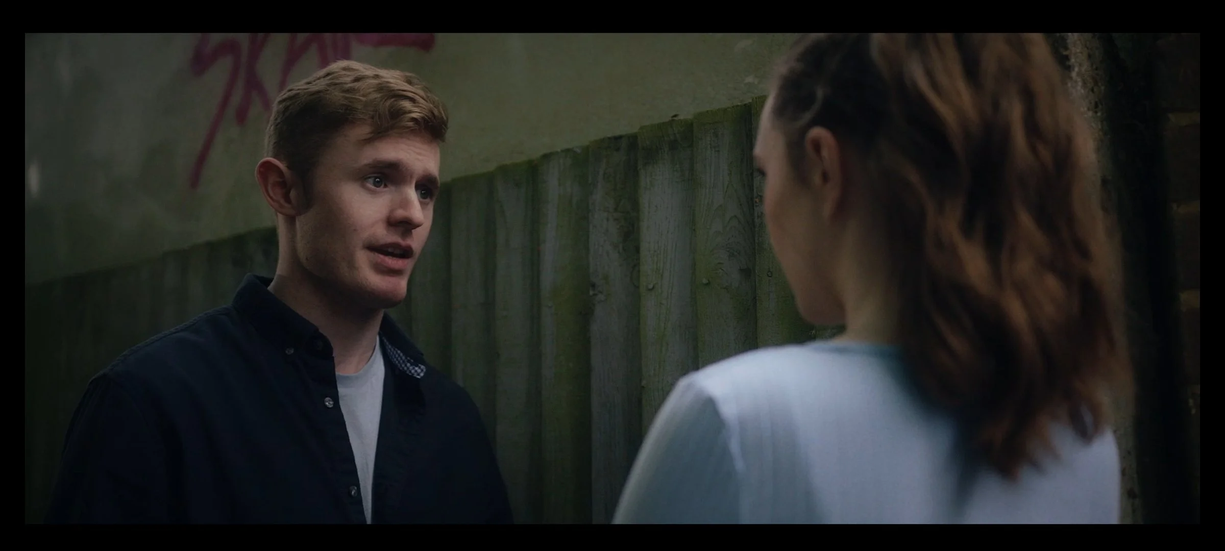A young man with red hair talking to a young woman with brown hair near a wooden fence in an outdoor setting.