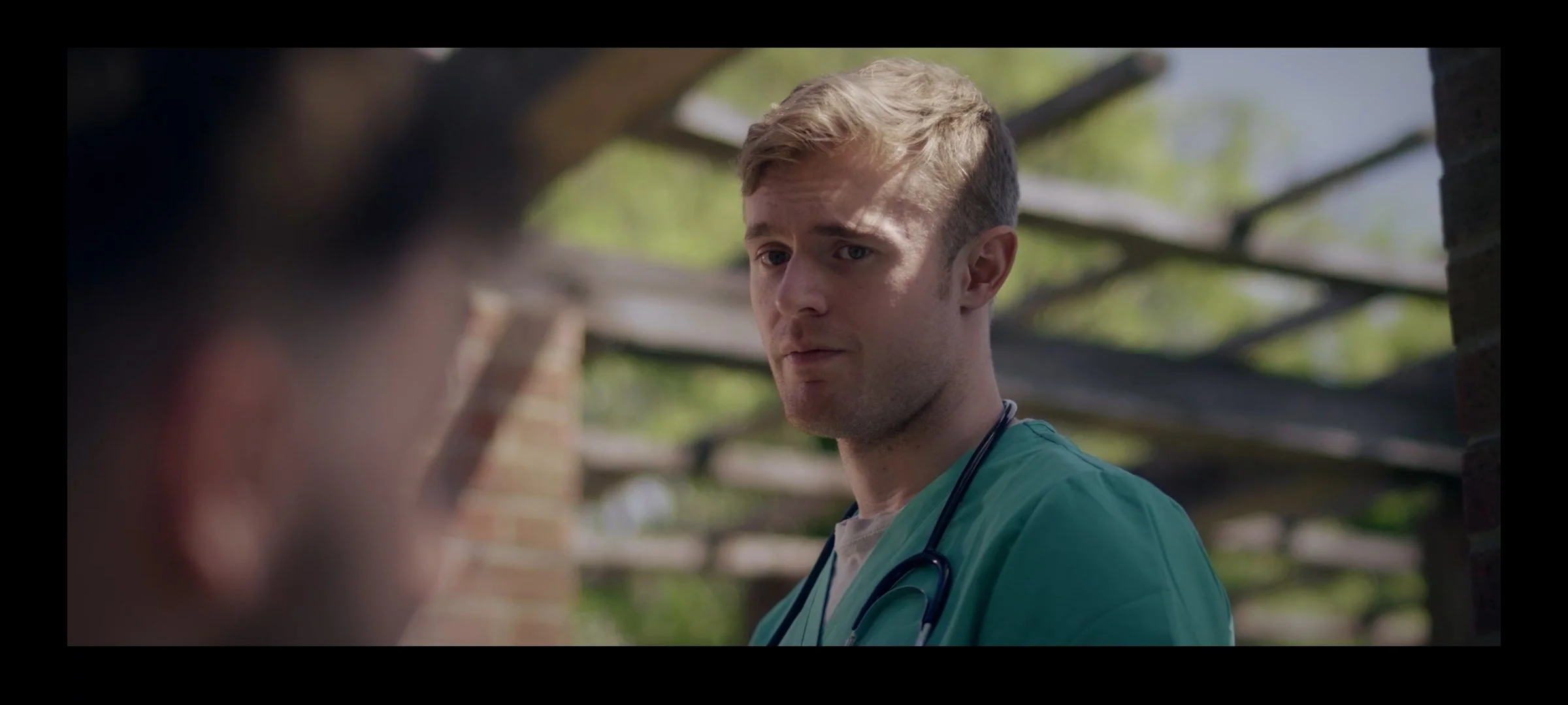 A young male surgeon with a stethoscope around his neck looks to the side at his patient, and former classmate, outdoors.