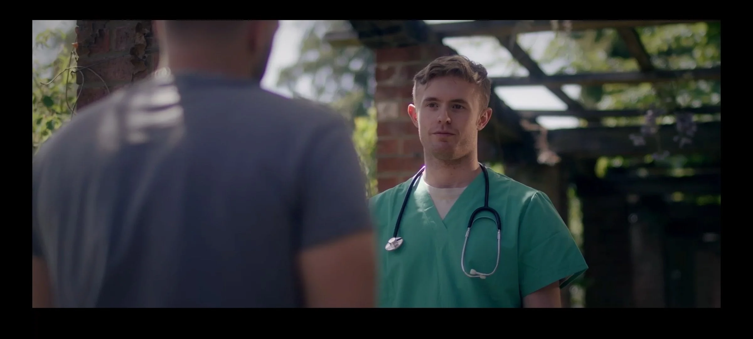 A young man wearing green medical scrubs and a stethoscope around his neck is looking at another person whose back is turned to the camera. The setting appears to be outdoors near a rustic, possibly abandoned, building with a wooden and brick structure and trees in the background.