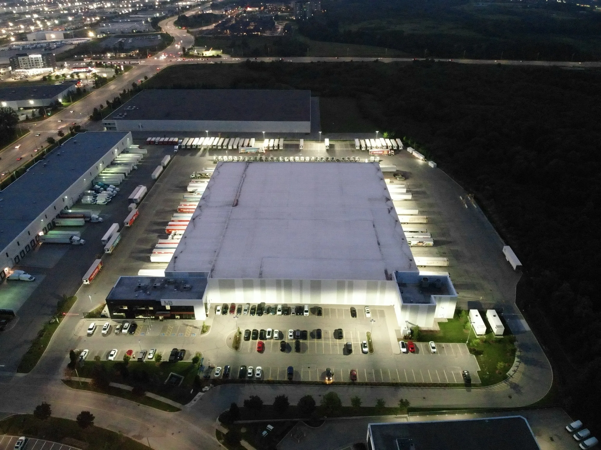 Aerial view of an illuminated warehouse complex with parking and loading docks at night.