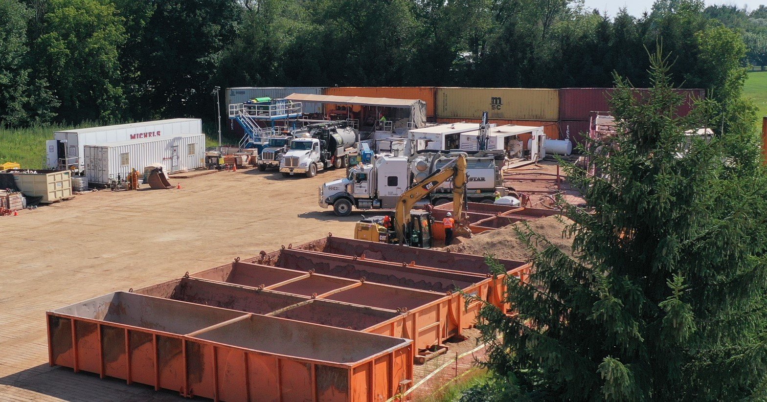 Industrial site with trucks, construction equipment, containers, and workers on a dirt lot surrounded by trees.