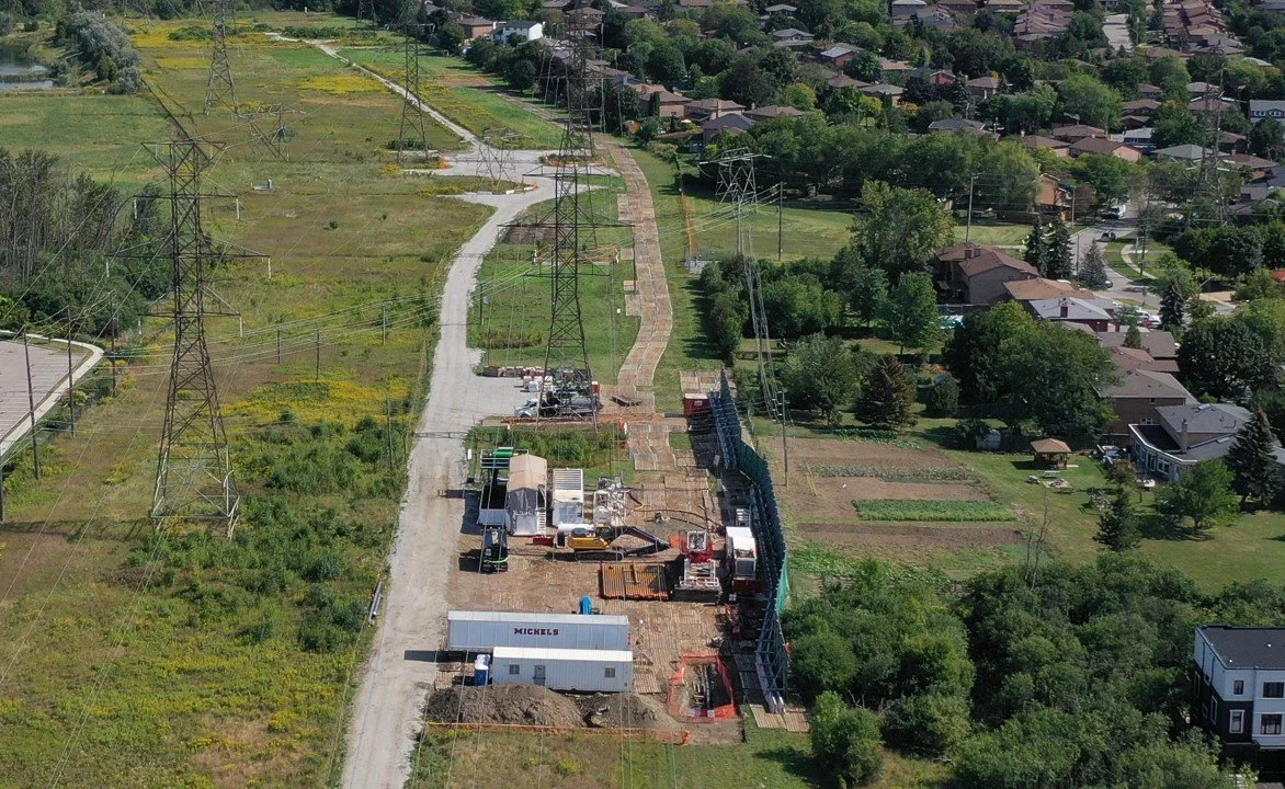 Aerial view of a construction site with heavy machinery and materials along a pathway lined with electricity pylons, surrounded by greenery and adjacent residential area.