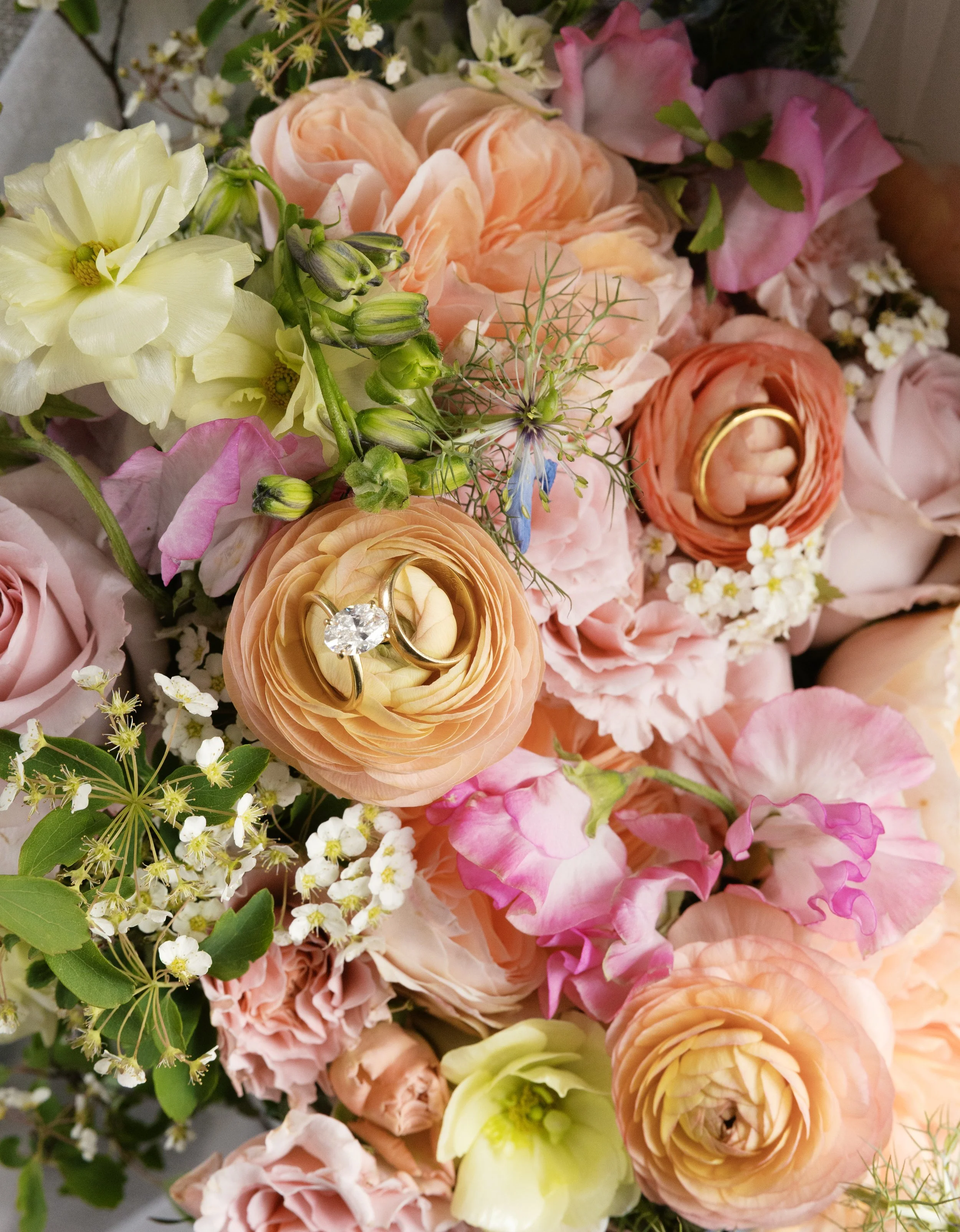 A close-up of a bouquet of assorted flowers, including pink and peach roses, white and yellow lisianthus, tiny white and yellow flowers, and greenery with delicate sprigs.