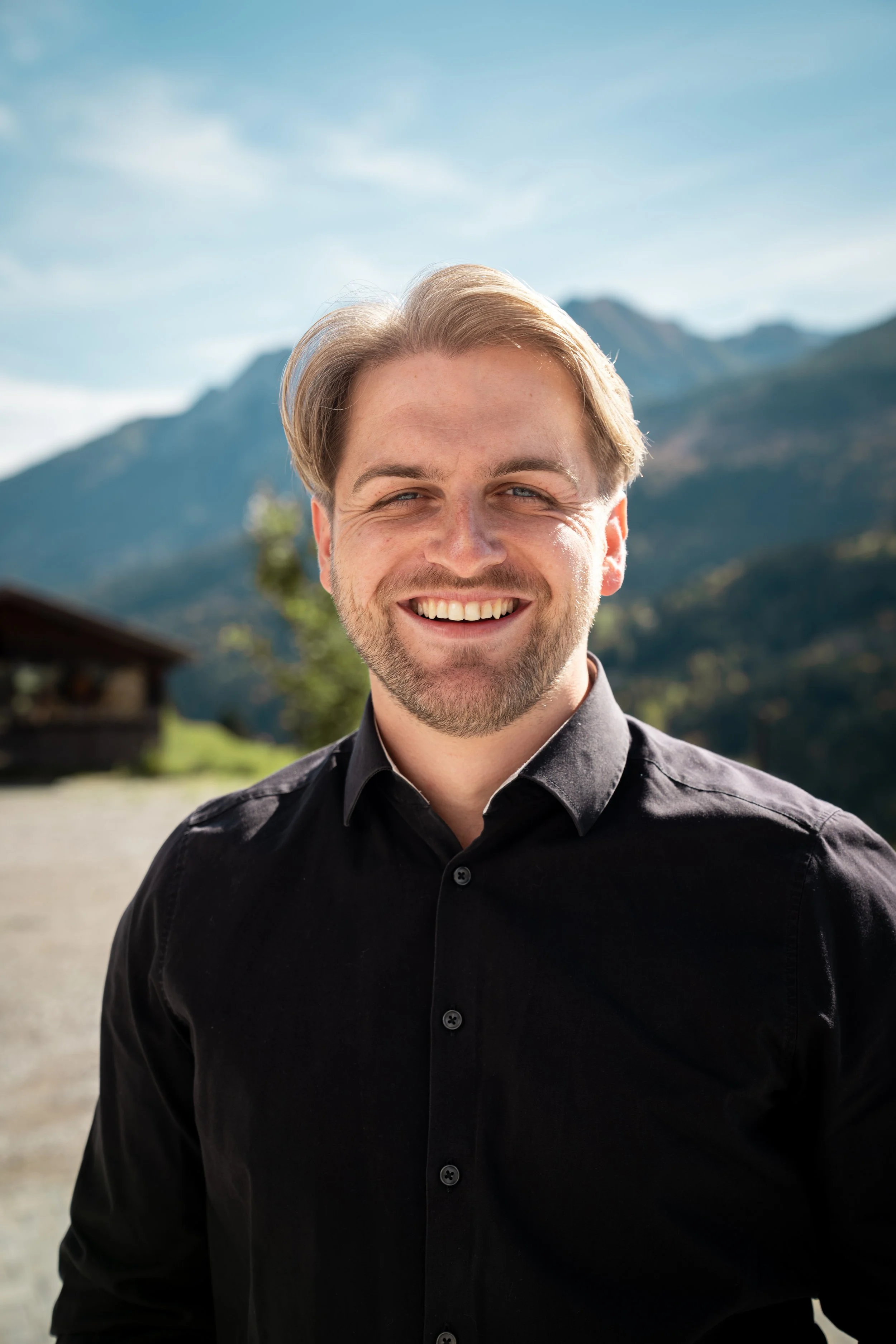 A smiling man with light brown hair and a beard, wearing a black shirt, outdoors with mountains and a cloudy sky in the background.
